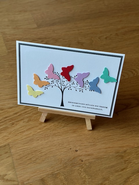Zu sehen ist eine selbst gebastelte Trauerkarte. An einem gestempelten Baum fliegen scheinbar sieben bunte Schmetterlinge vorbei. Einzig ein schwarzer Rand hinter dem Bild greift das klassische Layout einer Trauerkarte auf.
