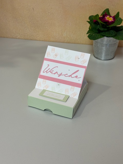 Zu sehen ist eine kleine Schachtel in grün. Oben auf ist eine Karte mit dem Wort "Wünsche" drauf zu sehen. Diese kann man wie eine Easelcard aufstellen. Klappt mann die Box auf. Erscheint die Rückansicht von einem Auto mit grüßen zur Hochzeit.