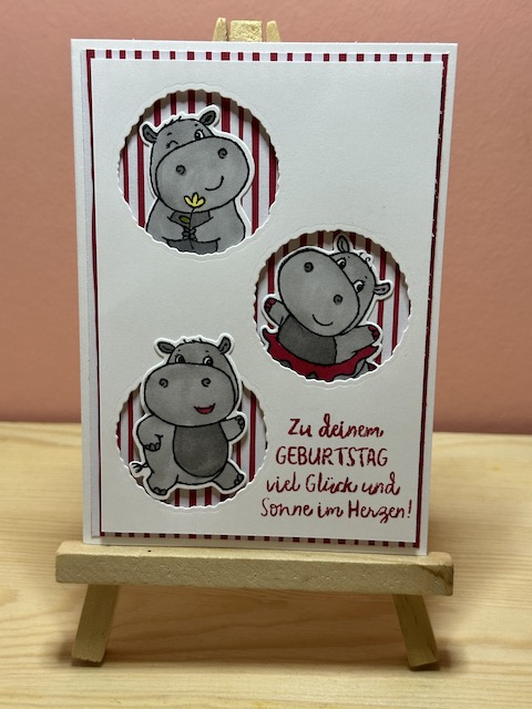 Zu sehen ist eine handmade Geburtstagskarte von Kerstins Kartenwerkstatt. Die Karte hat im weißen Layer drei Kreise ausgestanzt, aus denen jeweils ein Nilpferd mit einem lustigen Gesicht herausschaut. Hinter dem Nilpferd kann man gestreiftes Designpapier in weiß rot bzw. weiß grün sehen. Auf der Karte steht unten in der Ecke der Spruch: Zu deinem Geburtstag viel Glück und Sonne im Herzen