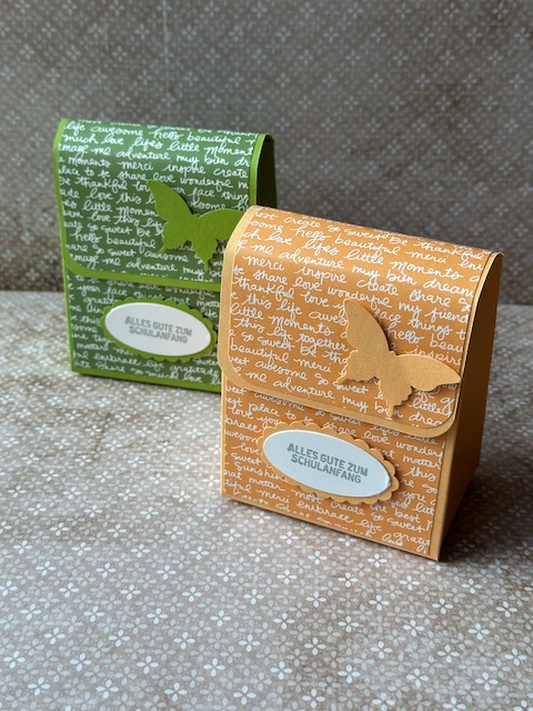 Zu sehen ist eine kleine selbstgebastelte Schultasche aus Papier. Der bunte Farbkarton ist mit Designpapier beklebt, dass mit Schrift bedruckt ist. Auf der Schultasche steht: Alles gute zum Schulanfang und ein Schmetterling ist auf der Klappe zu sehen. 