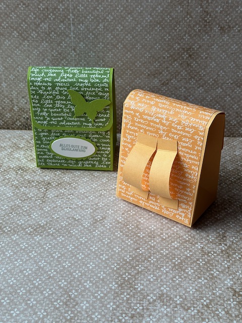 Zu sehen ist eine kleine selbstgebastelte Schultasche aus Papier. Der bunte Farbkarton ist mit Designpapier beklebt, dass mit Schrift bedruckt ist. Auf der Schultasche steht: Alles gute zum Schulanfang und ein Schmetterling ist auf der Klappe zu sehen. 