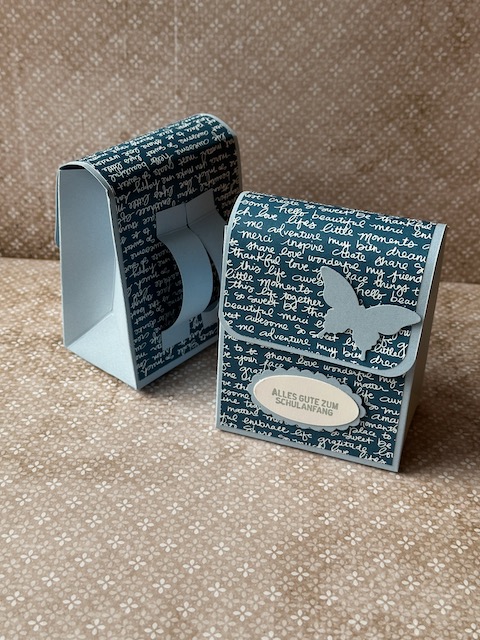 Zu sehen ist eine kleine selbstgebastelte Schultasche aus Papier. Der bunte Farbkarton ist mit Designpapier beklebt, dass mit Schrift bedruckt ist. Auf der Schultasche steht: Alles gute zum Schulanfang und ein Schmetterling ist auf der Klappe zu sehen. 