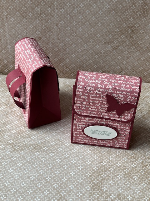Zu sehen ist eine kleine selbstgebastelte Schultasche aus Papier. Der bunte Farbkarton ist mit Designpapier beklebt, dass mit Schrift bedruckt ist. Auf der Schultasche steht: Alles gute zum Schulanfang und ein Schmetterling ist auf der Klappe zu sehen.