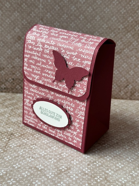 Zu sehen ist eine kleine selbstgebastelte Schultasche aus Papier. Der bunte Farbkarton ist mit Designpapier beklebt, dass mit Schrift bedruckt ist. Auf der Schultasche steht: Alles gute zum Schulanfang und ein Schmetterling ist auf der Klappe zu sehen. 