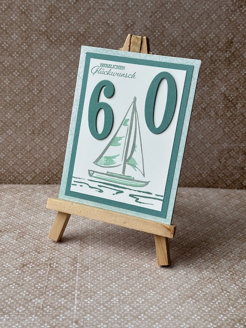 Zu sehen ist eine Geburtstagskarte in grün Tönen mit einem gestempelten Segelboot und Herzlichen Glückwunsch drauf. Zum 60. Geburtstag