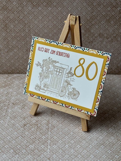 Zu sehen ist eine gebastelte Geburtstagskarte. Vorne drauf ist eine Tür samt dekorierten Rahmen und angelehntem Fahrrad zu sehen. Oben drüber steht: Alles gute zum Geburtstag. Hinterlegt ist das ganze durch einen Farbspiegel und Mosaik Designpapier. 