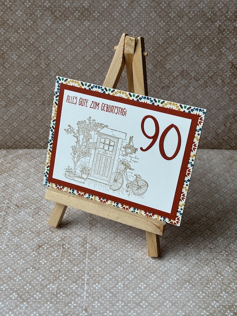 Zu sehen ist eine gebastelte Geburtstagskarte. Vorne drauf ist eine Tür samt dekorierten Rahmen und angelehntem Fahrrad zu sehen. Oben drüber steht: Alles gute zum Geburtstag. Hinterlegt ist das ganze durch einen Farbspiegel und Mosaik Designpapier. 