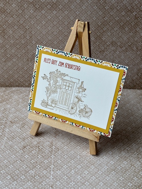 Zu sehen ist eine gebastelte Geburtstagskarte. Vorne drauf ist eine Tür samt dekorierten Rahmen und angelehntem Fahrrad zu sehen. Oben drüber steht: Alles gute zum Geburtstag. Hinterlegt ist das ganze durch einen Farbspiegel und Mosaik Designpapier. 