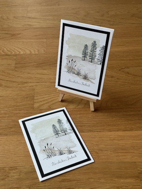 Zu sehen ist eine selbst gemachte Trauerkarte. Auf einer weißen Grundkarte ist ein schwarze Rahmen zu sehen und darauf ein bestempeltes Papier. Ein Hintergrundstempel sorgt für dezente Farbe. Für die Tiefe sorgen hinten ein paar Bäume und Vorne sind Grashalme und Schilf abgestempelt. Die Karte wird ergänzt mit dem Spruch: Herzliches Beileid