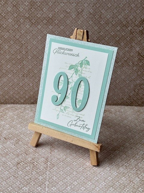 Zu sehen ist eine selbstgemachte Geburtstagskarte. Auf einer Grundkarte in einem Mintgrün sind zwei Lagen Papier geklebt. Die untere ebenfalls in einem grünlichen Ton die obere in weiß. Auf der Oberen ist ein dezenter Hintergrund gestempelt. Ebenfall ist Herzlichen Glückwunsch auf die Karte gestempelt. Zusätzlich sind ausgestanzte Ziffern aufgeklebt. Hier die 90