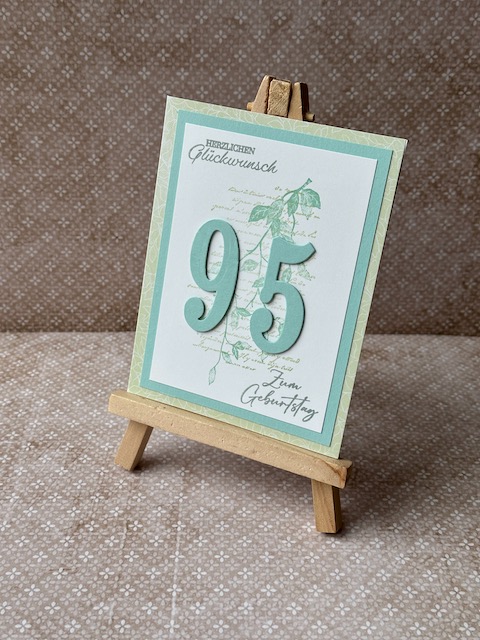 Zu sehen ist eine selbstgemachte Geburtstagskarte. Auf einer Grundkarte in einem Mintgrün sind zwei Lagen Papier geklebt. Die untere ebenfalls in einem grünlichen Ton die obere in weiß. Auf der Oberen ist ein dezenter Hintergrund gestempelt. Ebenfall ist Herzlichen Glückwunsch auf die Karte gestempelt. Zusätzlich sind ausgestanzte Ziffern aufgeklebt. Hier die 95