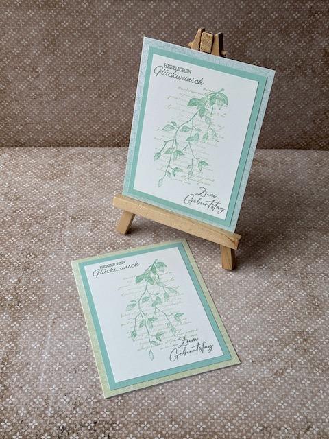 Zu sehen ist eine selbstgemachte Geburtstagskarte. Auf einer Grundkarte in einem Mintgrün sind zwei Lagen Papier geklebt. Die untere ebenfalls in einem grünlichen Ton die obere in weiß. Auf der Oberen ist ein dezenter Hintergrund gestempelt. Ebenfalls ist Herzlichen Glückwunsch auf die Karte gestempelt.