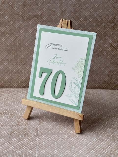 Zu sehen ist eine selbstgemachte Geburtstagskarte. Auf einer Grundkarte in einem Mintgrün sind zwei Lagen Papier geklebt. Die untere ebenfalls in einem grünlichen Ton die obere in weiß. Auf der Oberen ist ein dezenter Hintergrund gestempelt. Ebenfall ist Herzlichen Glückwunsch auf die Karte gestempelt. Zusätzlich sind ausgestanzte Ziffern aufgeklebt. Hier die 70