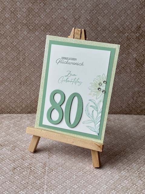 Zu sehen ist eine selbstgemachte Geburtstagskarte. Auf einer Grundkarte in einem Mintgrün sind zwei Lagen Papier geklebt. Die untere ebenfalls in einem grünlichen Ton die obere in weiß. Auf der Oberen ist ein dezenter Hintergrund gestempelt. Ebenfall ist Herzlichen Glückwunsch auf die Karte gestempelt. Zusätzlich sind ausgestanzte Ziffern aufgeklebt. Hier die 80