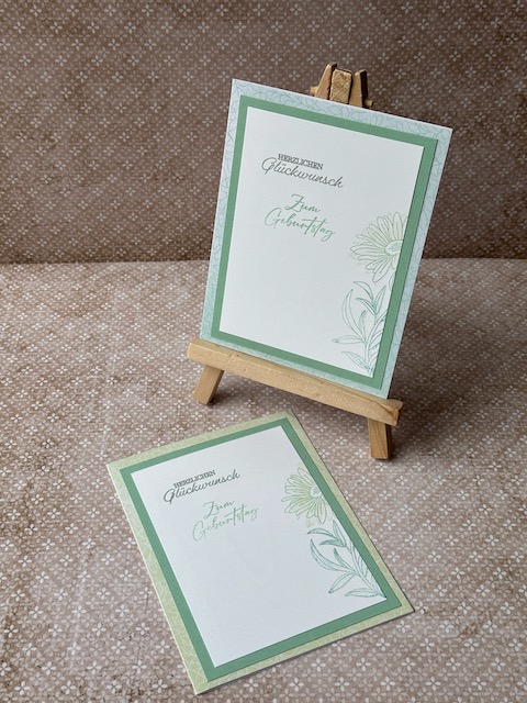 Zu sehen ist eine selbstgemachte Geburtstagskarte. Auf einer Grundkarte in einem Mintgrün sind zwei Lagen Papier geklebt. Die untere ebenfalls in einem grünlichen Ton die obere in weiß. Auf der Oberen ist ein dezenter Hintergrund gestempelt. Ebenfalls ist Herzlichen Glückwunsch auf die Karte gestempelt.