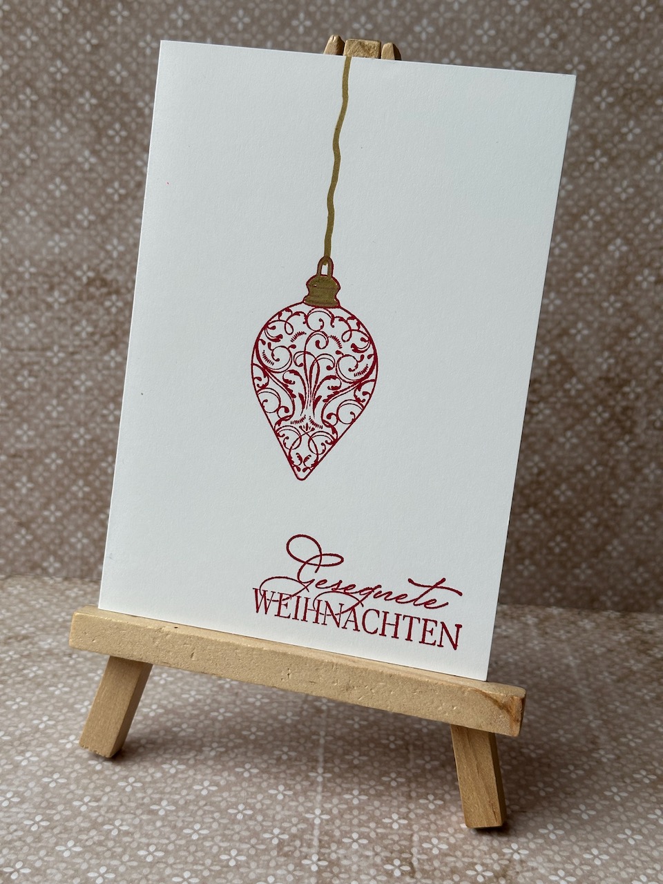 Zu sehen ist eine minimalistische Weihnachstkarte. Auf einer weißen Grundkarte ist mittig in rot Anhänger in Zapfenform gestempelt. In goldener feiner Linie ist bis nach oben an den Rand ein Linie gemalt, die andeutet, dass der Zapfen hängt. Ergänz wird das Bild durch einen weihnachtlichen Gruß. 