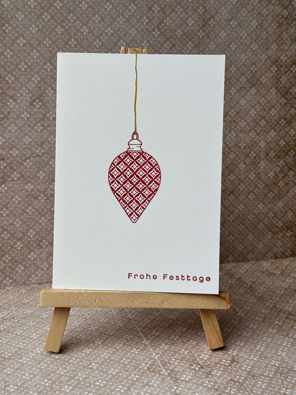 Zu sehen ist eine minimalistische Weihnachstkarte. Auf einer weißen Grundkarte ist mittig in rot Anhänger in Zapfenform gestempelt. In goldener feiner Linie ist bis nach oben an den Rand ein Linie gemalt, die andeutet, dass der Zapfen hängt. Ergänz wird das Bild durch einen weihnachtlichen Gruß. 