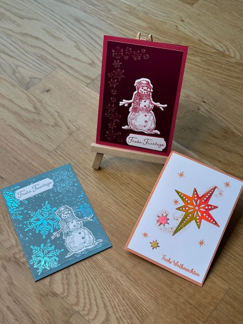 Selbstgebastelte Weihnachtskarten mit metallischen Akzenten auf Papier. In blau, orange und rot. Jeweils mit Schneemann oder Stern und einem besonderen Glitzern, wenn man die Karte im Licht bewegt.